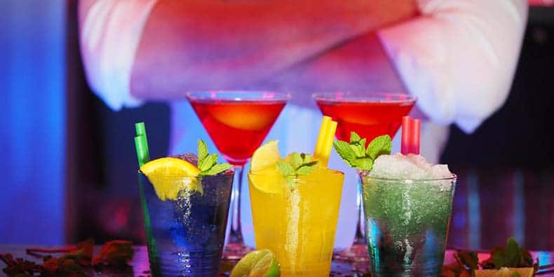 Bartender standing at bar behind several different colourful mixed drinks