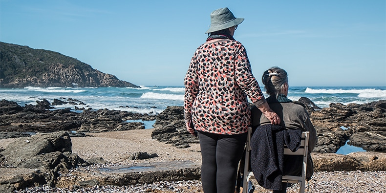 Senior woman sitting in a chair at beach with companion behind her as they look out at ocean