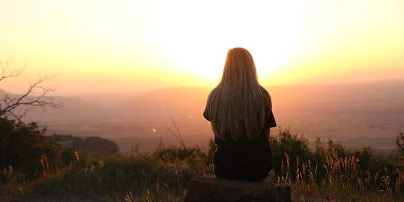Young woman sitting in field overlooking valley at sunset; introspective