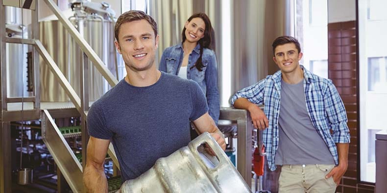 Young man holding keg with these colleagues behind him in the factory