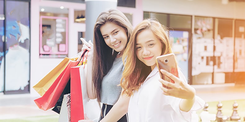 Young Millennial girls taking smart phone selfie and holding bag in department store. Soft flare filter.