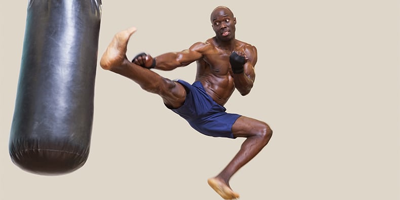 Full length of a shirtless muscular boxer kicking punching bag over white background