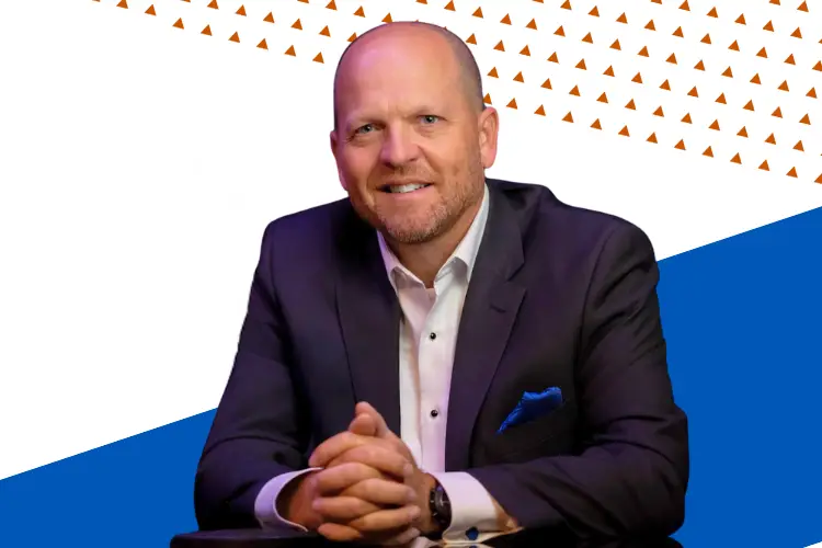 A professionally dressed man with a bald head and trimmed beard smiles while seated with his hands clasped on a table. He is wearing a dark suit, white shirt, and a blue pocket square. The background features a bold geometric design with white, orange triangle patterns, and blue sections.