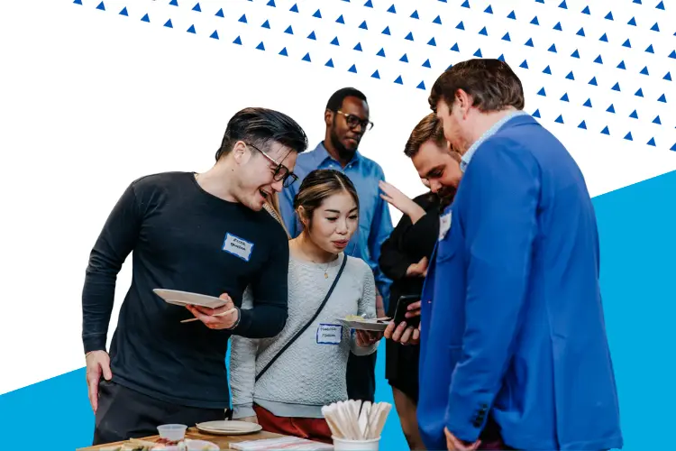 A group of professionally dressed people gather around a table at a networking event, smiling and looking at a phone. They wear name tags and hold small plates of food, with a modern blue and white geometric background featuring blue triangles.