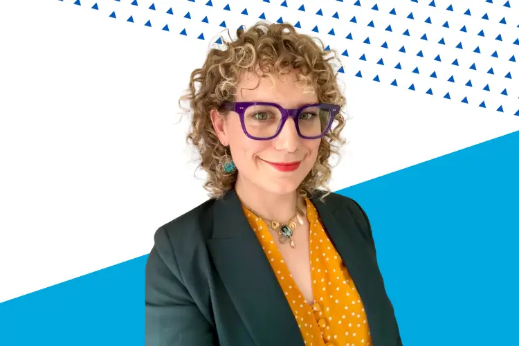 A person with curly blond hair, wearing bold purple glasses, a dark teal blazer, and a yellow polka-dot blouse smiles at the camera. The background features a white and blue geometric design with small blue triangles.